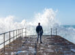 Business man on harbour looking out to sea with wave crashing against wall.