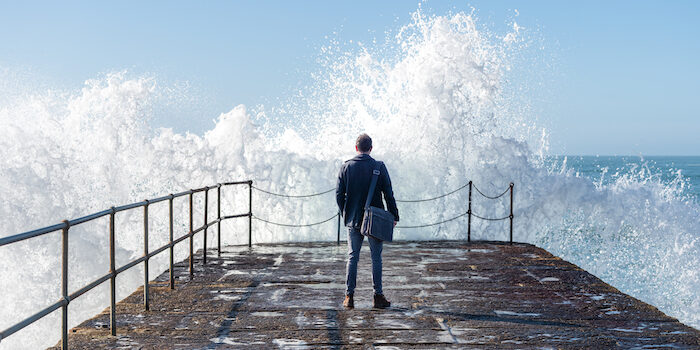 Business man on harbour looking out to sea with wave crashing against wall.