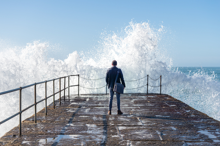 Business man on harbour looking out to sea with wave crashing against wall.